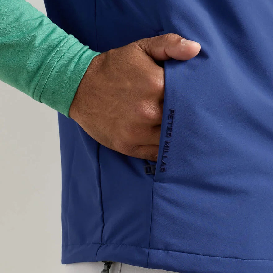 A person wearing a light green long-sleeve shirt and a blue Peter Millar Boeing Men’s Dunes Performance Vest with performance fabric puts their hand in the pocket, showing the BOEING logo near the pocket.