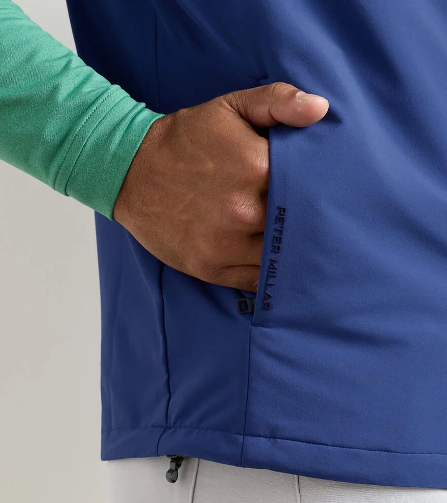 A person wearing a light green long-sleeve shirt and a blue Peter Millar Boeing Men’s Dunes Performance Vest with performance fabric puts their hand in the pocket, showing the BOEING logo near the pocket.