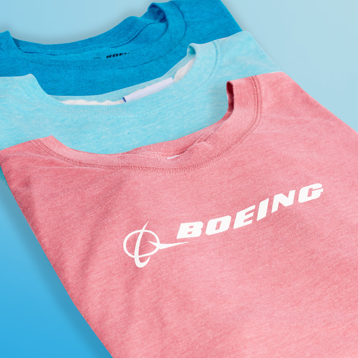  close-up of three t-shirts in different colors (blue and pink) featuring the Boeing logo, arranged neatly against a light blue background.