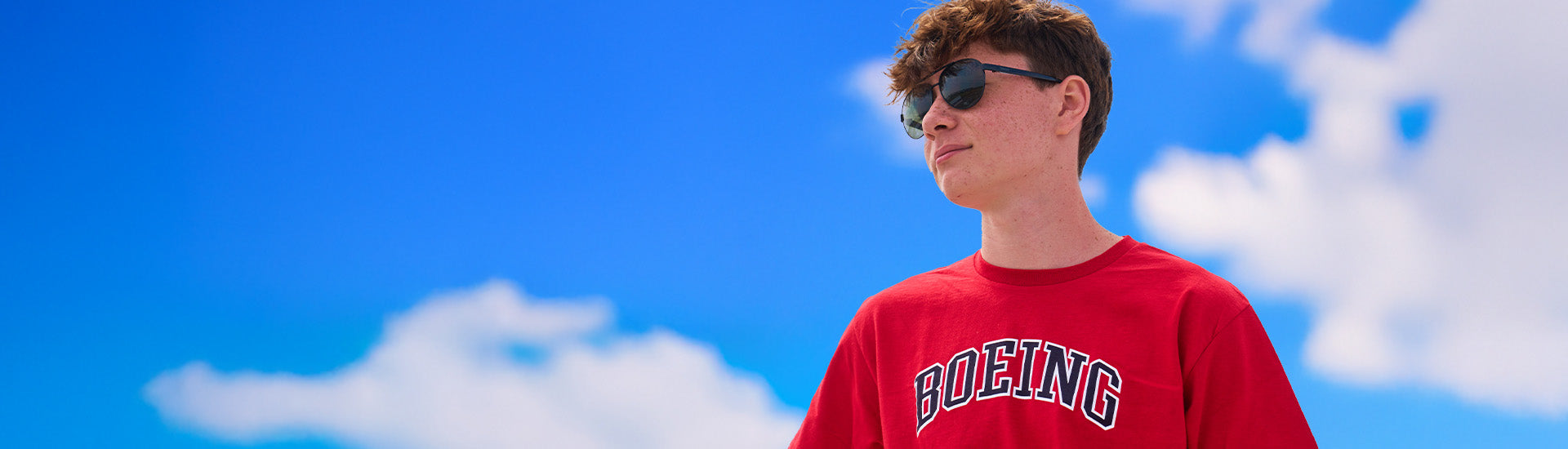 Desktop banner featuring a young man wearing a red t-shirt with "Boeing" printed on it, posed against a blue sky background.