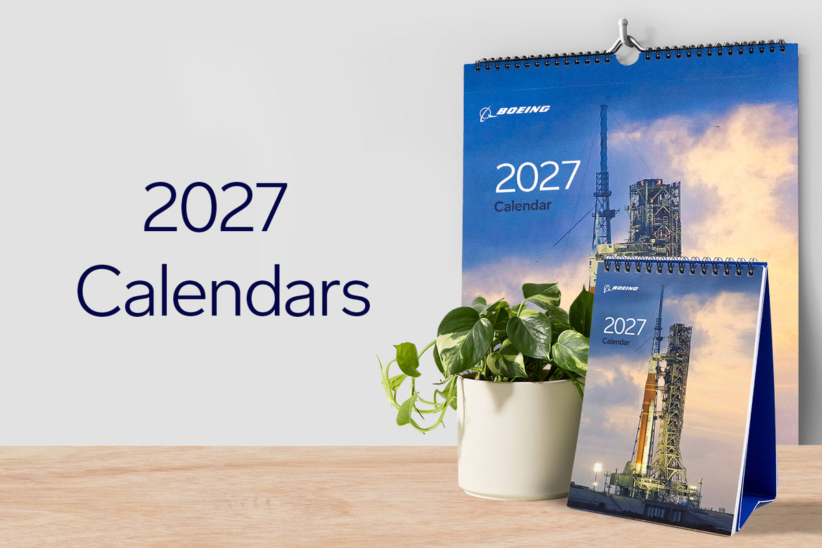 A 2027 Boeing wall calendar and desk calendar featuring a rocket launch pad, displayed with a potted plant on a wooden surface.