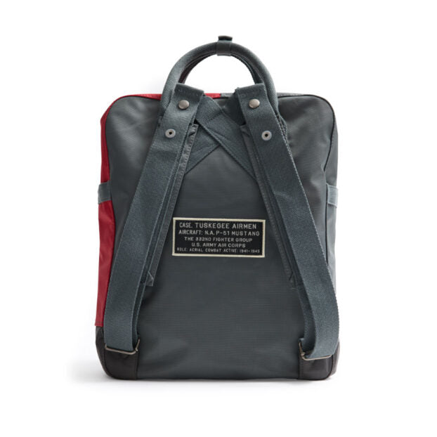 Back view of a grey and red Red Canoe Boeing Tuskegee Red Tails Backpack made of durable nylon, featuring a rectangular patch with text detailing the history of the Tuskegee Airmen.