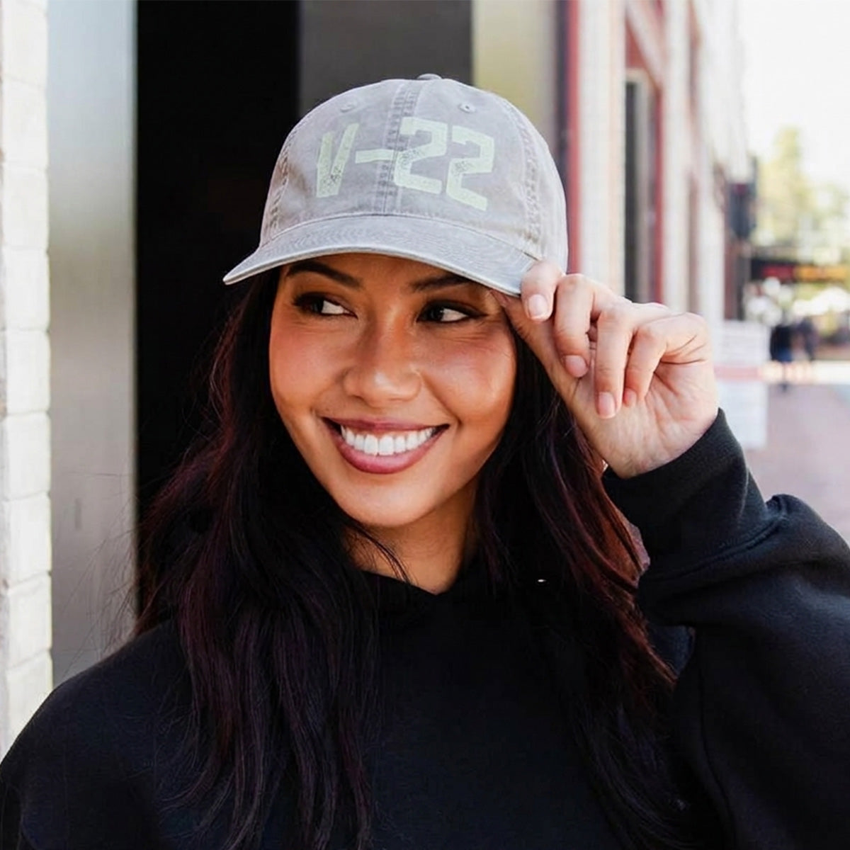 A smiling woman in a black hoodie adjusts a gray Boeing V-22 Osprey Graphic Hat by BOEING on a city street—an ideal accessory for aviation fans and Boeing supporters.