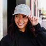 A smiling woman in a black hoodie adjusts a gray Boeing V-22 Osprey Graphic Hat by BOEING on a city street—an ideal accessory for aviation fans and Boeing supporters.