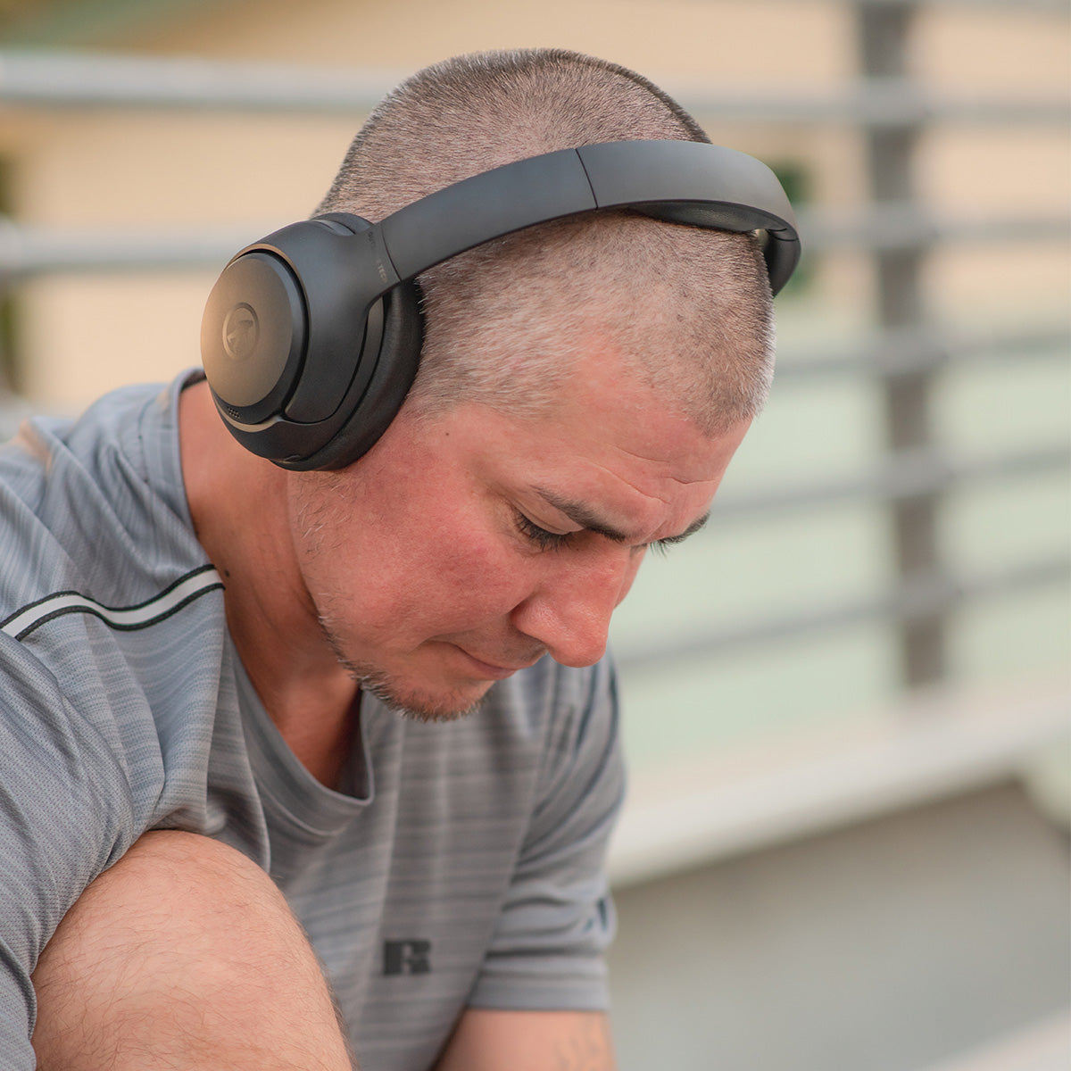 A man with short hair and a trimmed beard, wearing a gray shirt and BOEING Outdoor Tech Boeing Orbit ANC Headphones, sits outdoors looking down thoughtfully.