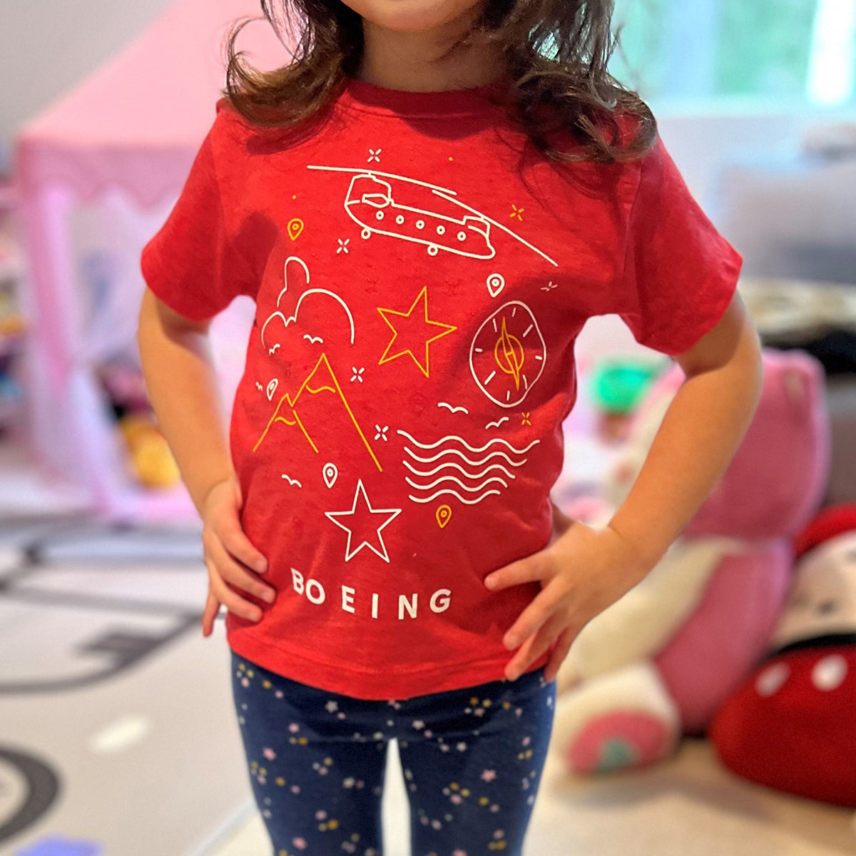 A toddler confidently stands in a playroom wearing a red Boeing Defense Doodle Toddler T-Shirt with plane and star graphics. With one hand on their hip, they are surrounded by a pink play tent and plush toys.