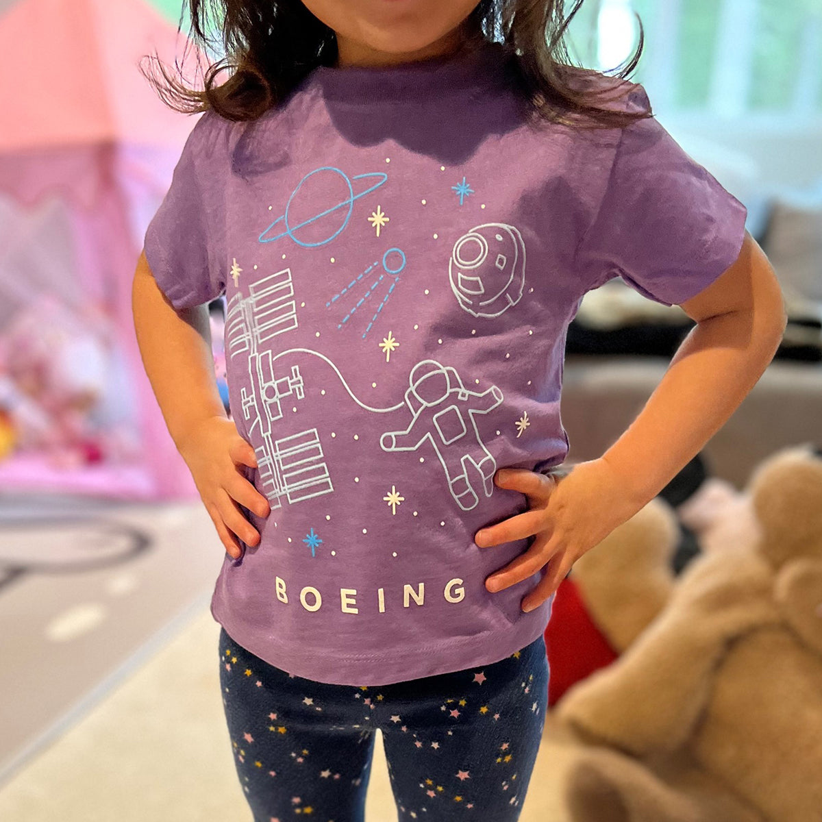 A toddler proudly wears the Boeing Space Doodle Toddler T-Shirt, featuring astronauts, planets, and spacecraft. Paired with star-patterned leggings, the outfit pops amid soft toys and a cozy play tent backdrop.