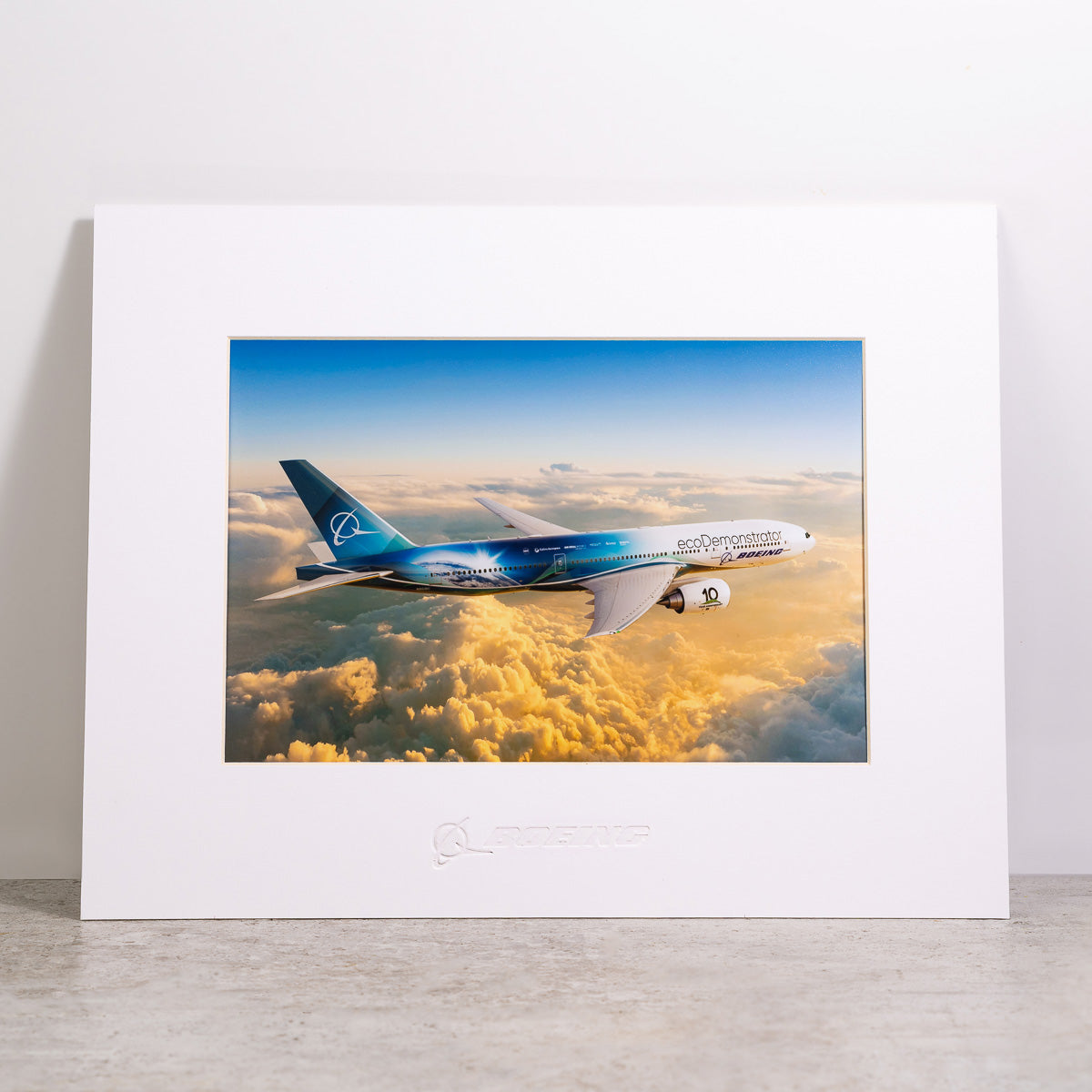 The Boeing Store’s Boeing 777-200ER ecoDemonstrator Matted Print – Small features a high-res image of a blue ecoDemonstrator jet flying above sunset clouds, with the Boeing logo embossed on the white mat.