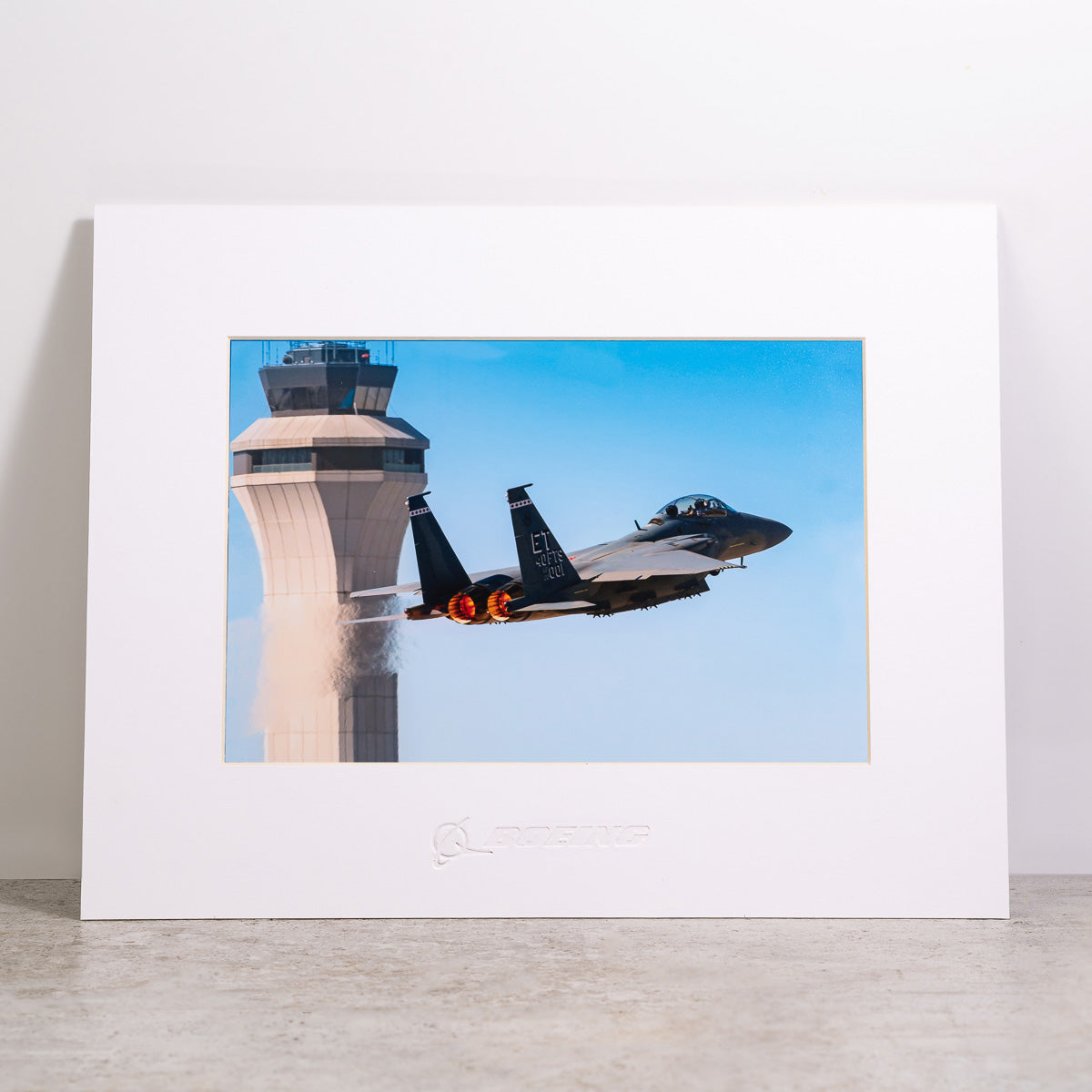 A Boeing F-15EX Eagle jet with afterburners engaged flies by a control tower in this vibrant matted print, featuring the Boeing logo. The scene is set against a solid, light-colored wall. Product: Boeing F-15EX Eagle Matted Print - Small by The Boeing Store.