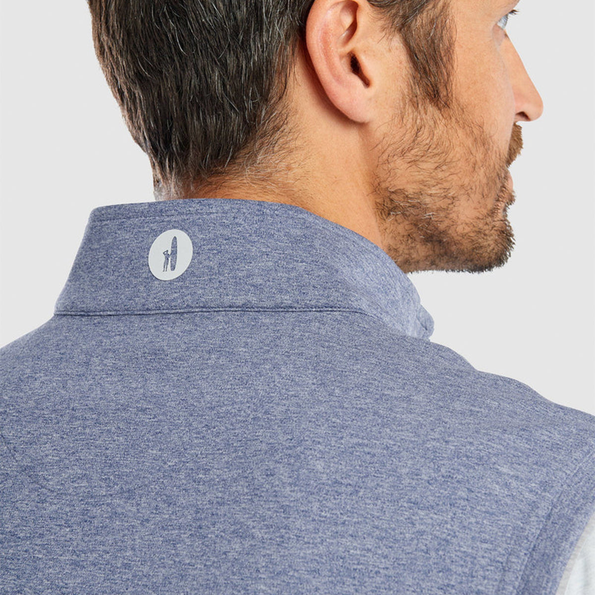 A man with short dark hair and a beard is seen from the back wearing The Boeing Stores johnnie-O Boeing Mens Denny Prep Performance Quarter-Zip Vest. The vest, adorned with a small white dog logo on the back collar, features moisture-wicking fabric.