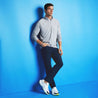 full body lifestyle image with male model standing against a blue wall.  Model wearing navy chino pants and shoes