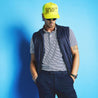 Product lifestyle image on a male model.  Vest is unzipped over a striped polo. Model is wearing a neon hat and standing against a blue wall