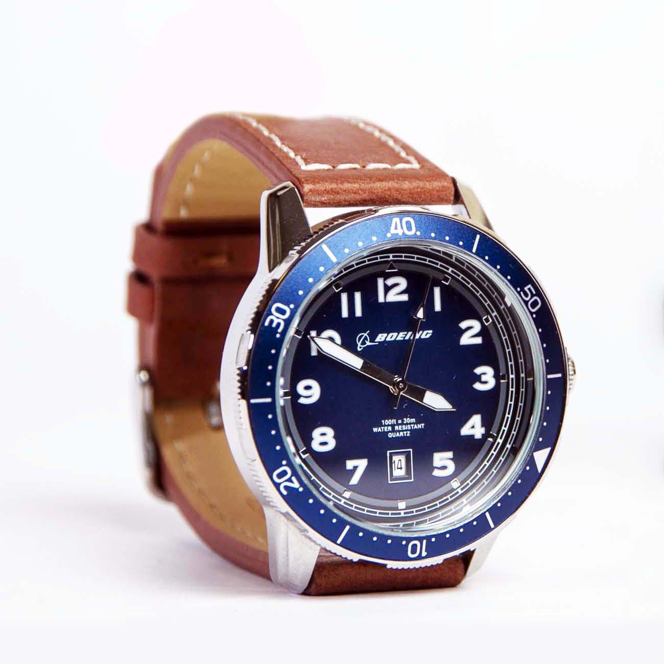 The Boeing Brown Leather Watch from The Boeing Store features a brown leather strap, stainless steel case, and blue bezel with white numerals. It showcases a blue dial with white hour markers, hands, and a date window at 6 oclock. Powered by Japanese movement for stylish precision.