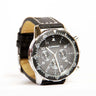 The Boeing Stores Boeing Black Leather Watch is a mens analog wristwatch boasting a Japanese movement, a stainless steel case with a black dial, three subdials, white markings including the number 60 at the top, and a tachymetric bezel for precision.