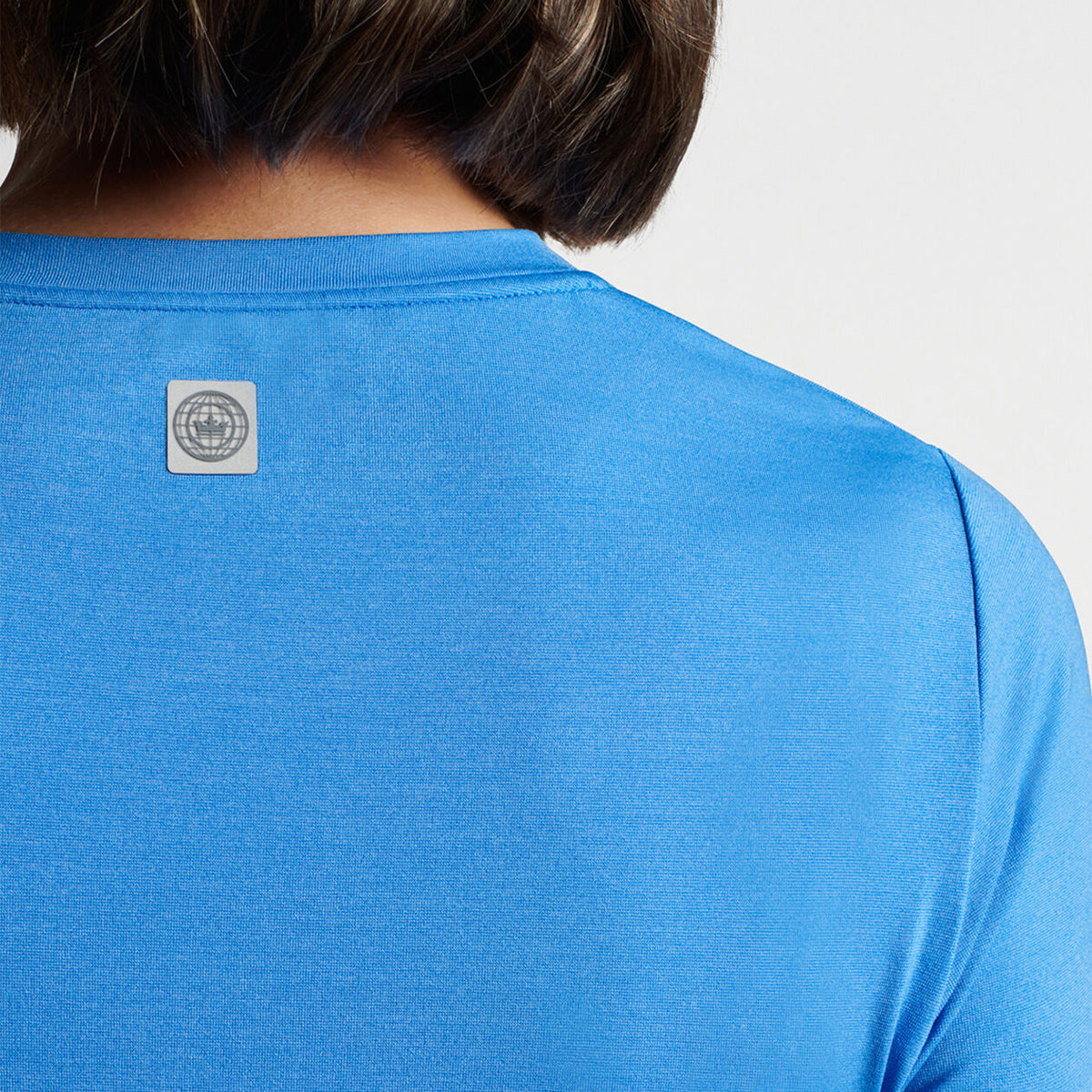 Close-up of a person in the Peter Millar Boeing Women’s Lunar Short-Sleeve Performance Tee, featuring a square logo with an abstract circular design on the upper back. Against a white backdrop, this blue tee from The Boeing Store offers UPF 50+ protection.