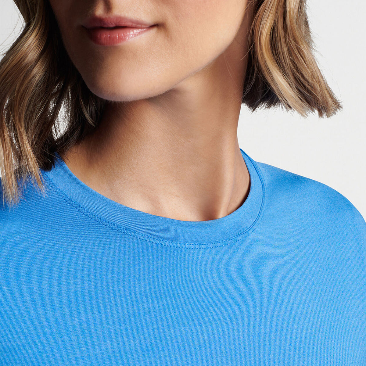 A person with short light brown hair wears a Peter Millar Boeing Womens Lunar Short-Sleeve Performance Tee in bright blue, highlighting its UPF 50+ sun protection. The focus is on the neckline and part of the face against a plain white background. Available at The Boeing Store.