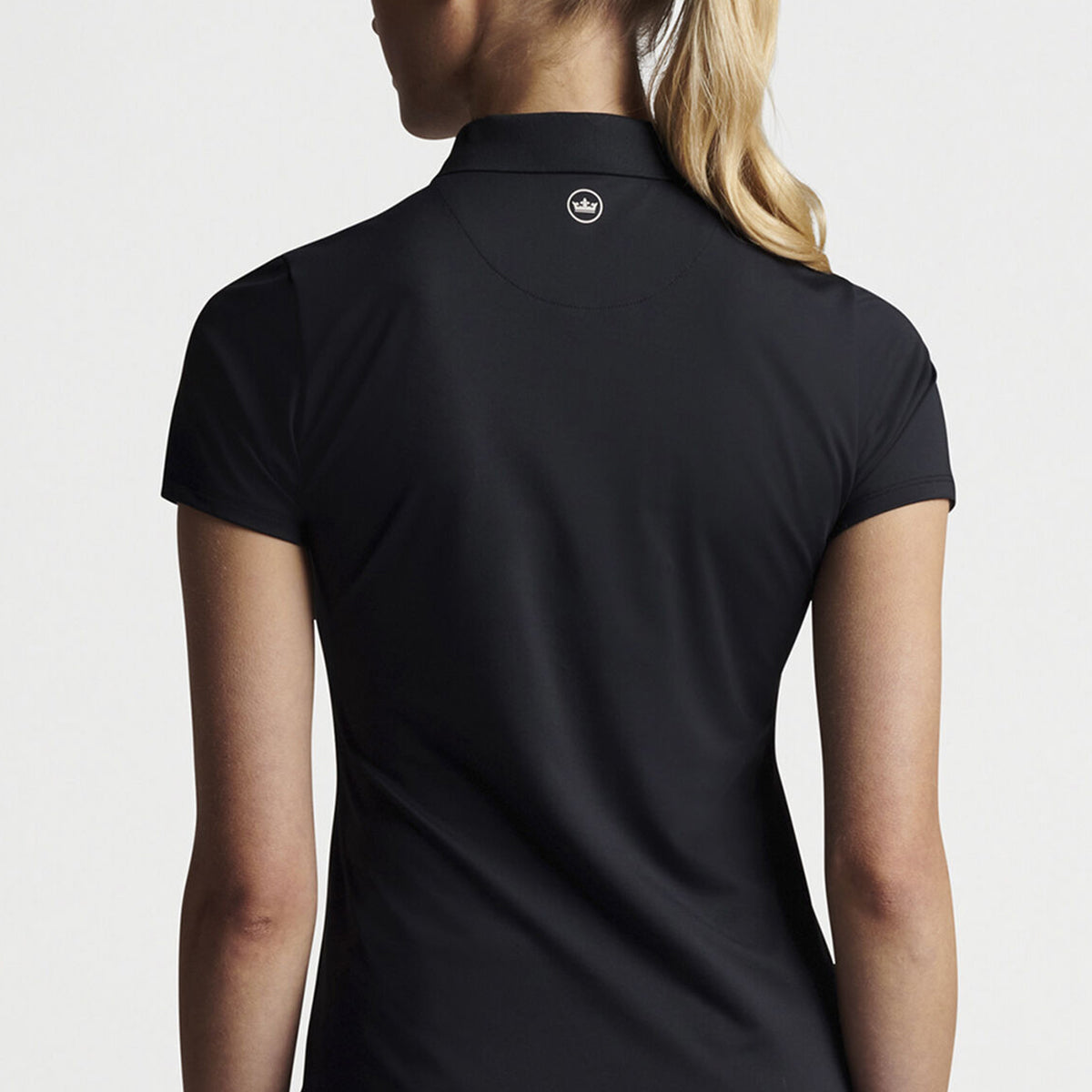 A woman with long, blonde hair tied back stands facing away, wearing a fitted Peter Millar Boeing Women’s Chrissie Short-Sleeve Polo. The sleek black performance polo from The Boeing Store features a small logo on the back collar and moisture-wicking fabric for comfort against a plain white backdrop.
