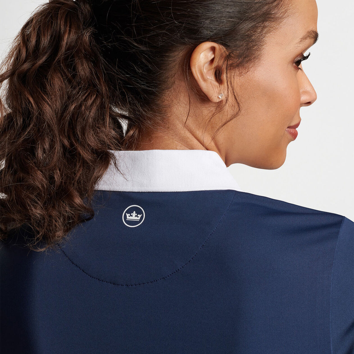 A woman with long, curly hair tied back is shown from the side and back wearing a navy Peter Millar Boeing Womens Chrissie Polo from The Boeing Store. It features performance fabric, a white collar, and a small crown emblem against a plain white background.