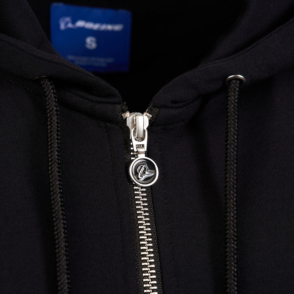 The Boeing Store’s Signature Logo Womens Black Short Waist Hoodie features a silver zipper with an embossed logo on the pull, an interior label showing size S, drawstrings beside the zipper, and stylish thumbholes.