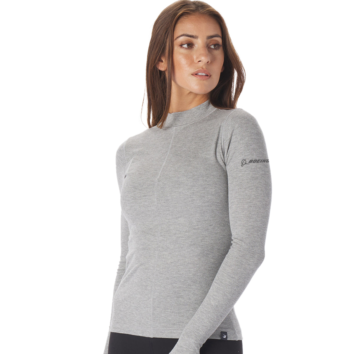 A woman models the elegant Glyder Boeing Womens Ruby Mock-Neck Long-Sleeve in light gray from The Boeing Store. With a small logo on the sleeve, she stands slightly turned, gazing thoughtfully over her shoulder with her hair down.