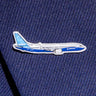 The Boeing Stores silver tone Boeing Illustrated 737 MAX Lapel Pin, featuring a blue and white airplane design, adds elegance to a dark blue fabric, like a suit jacket.