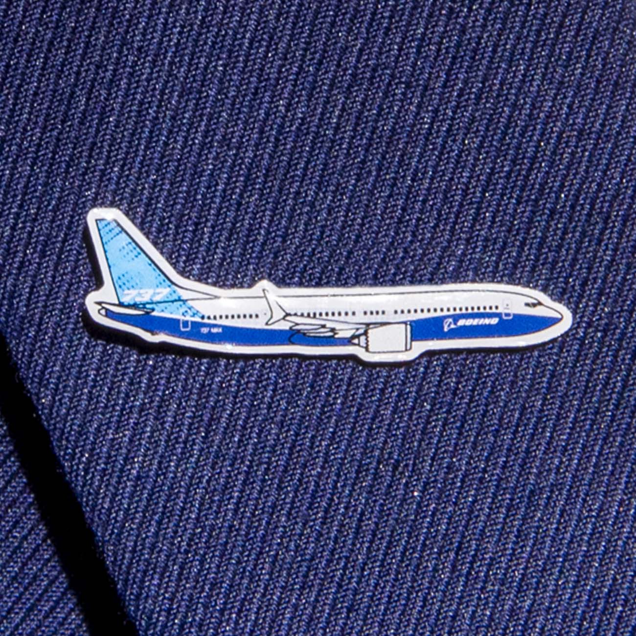 The Boeing Stores silver tone Boeing Illustrated 737 MAX Lapel Pin, featuring a blue and white airplane design, adds elegance to a dark blue fabric, like a suit jacket.