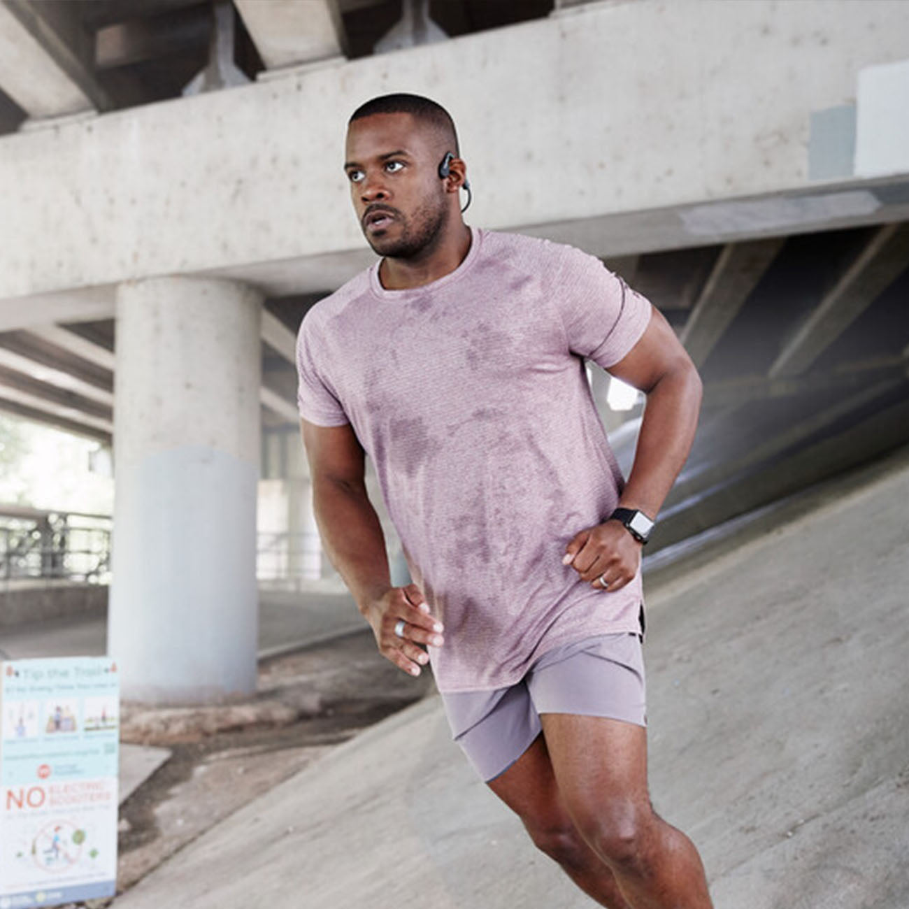 A man in a sweat-soaked pink shirt and shorts runs outdoors under a concrete structure. He sports a Shokz™ OpenMove Headset from The Boeing Store with bone conduction technology and a smartwatch. An unreadable safety sign looms in the background, but he stays focused, moving to his Bluetooth v5.0 playlist.
