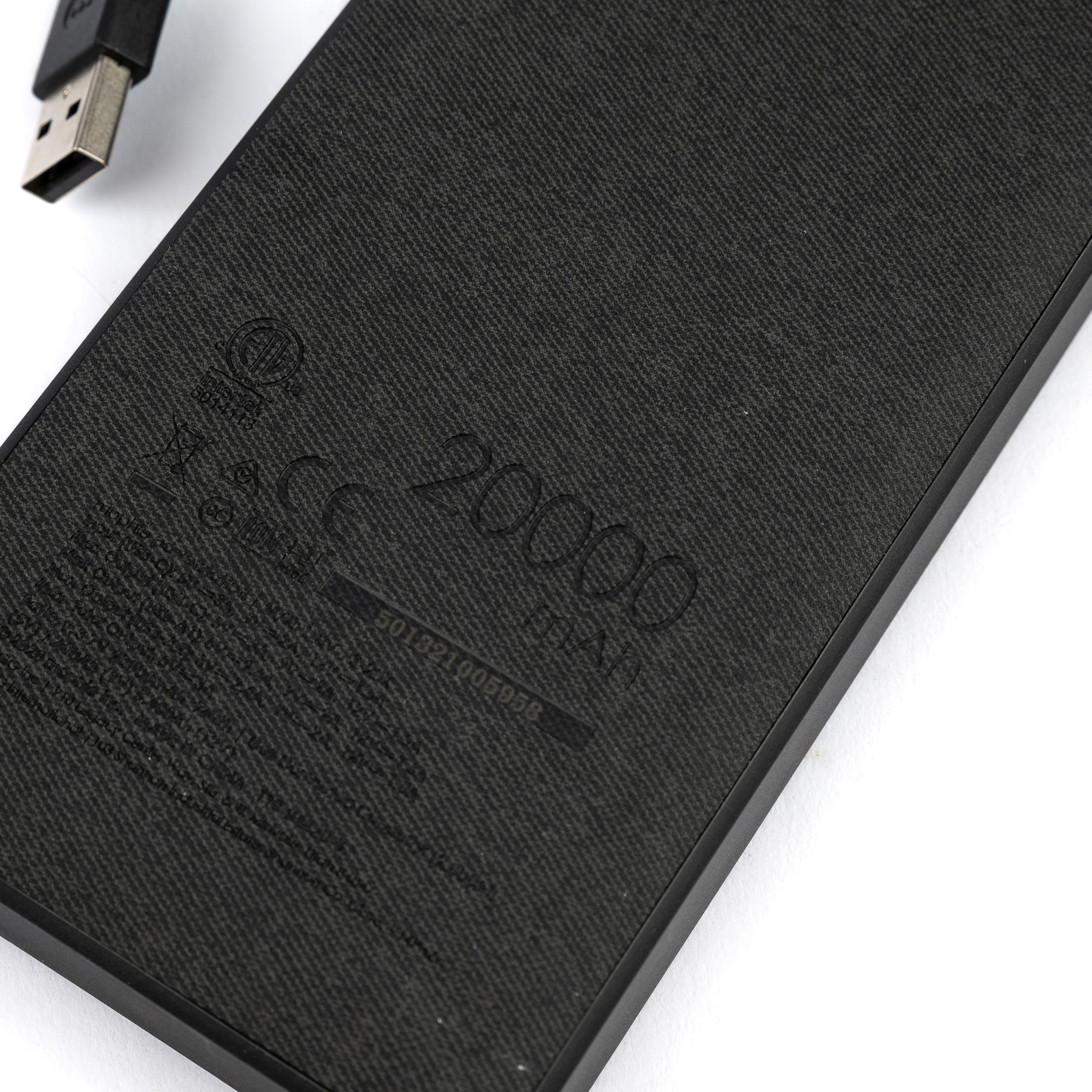 Close-up of the Mophie Boeing Powerstation XXL by The Boeing Store, featuring its impressive 20000 mAh capacity. A USB cable hovers above a port, while its textured fabric-like finish adds elegance. Faint symbols and serial numbers complete the sophisticated look.