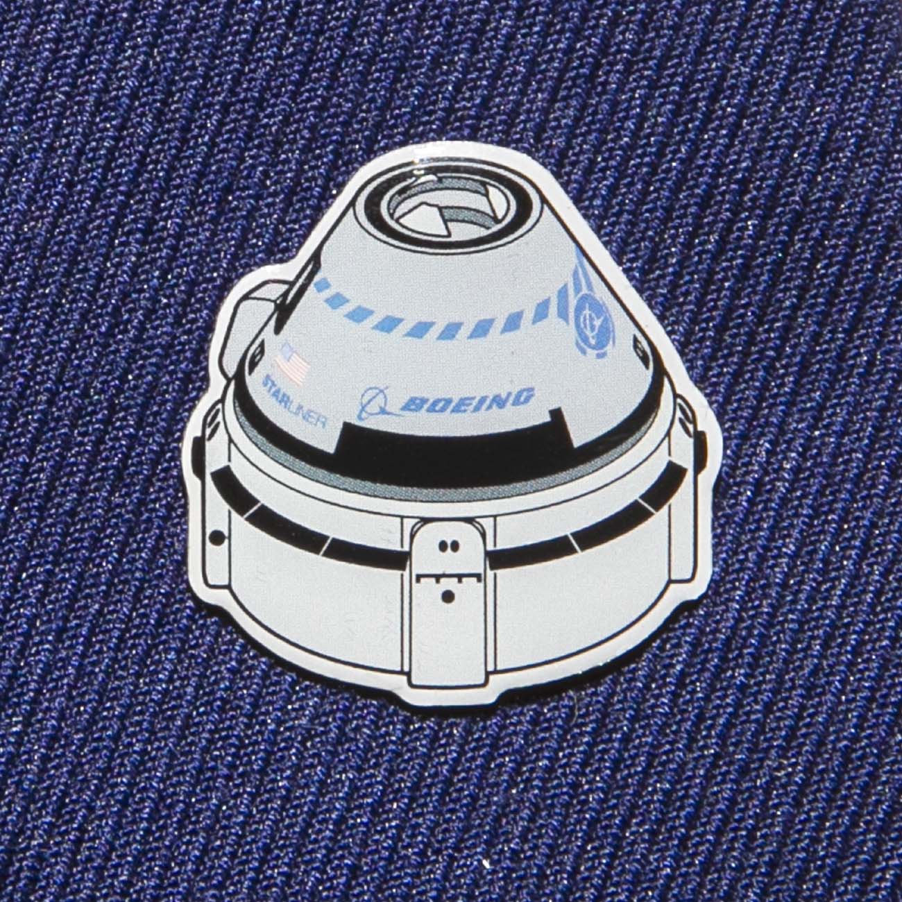A Boeing Illustrated Starliner Lapel Pin by The Boeing Store features a spacecraft design on blue fabric. It showcases an epoxy finish and small logos, including the Boeing logo, for a realistic touch.