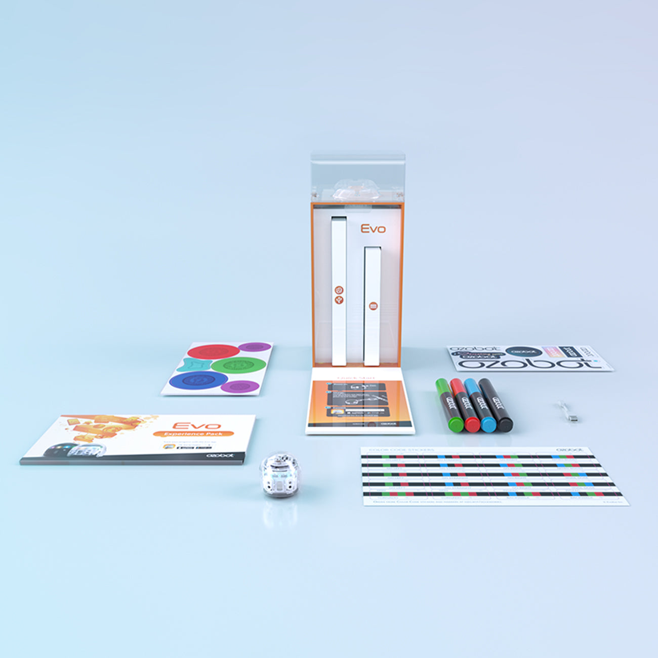 A tabletop displays an Ozobot Evo coding robot along with markers, color-coded stickers, and booklets featuring the same design. A box bears identical text and imagery beside a card with colored lines and a symbol sheet for OzoBlockly programming using the Evo app from The Boeing Store.