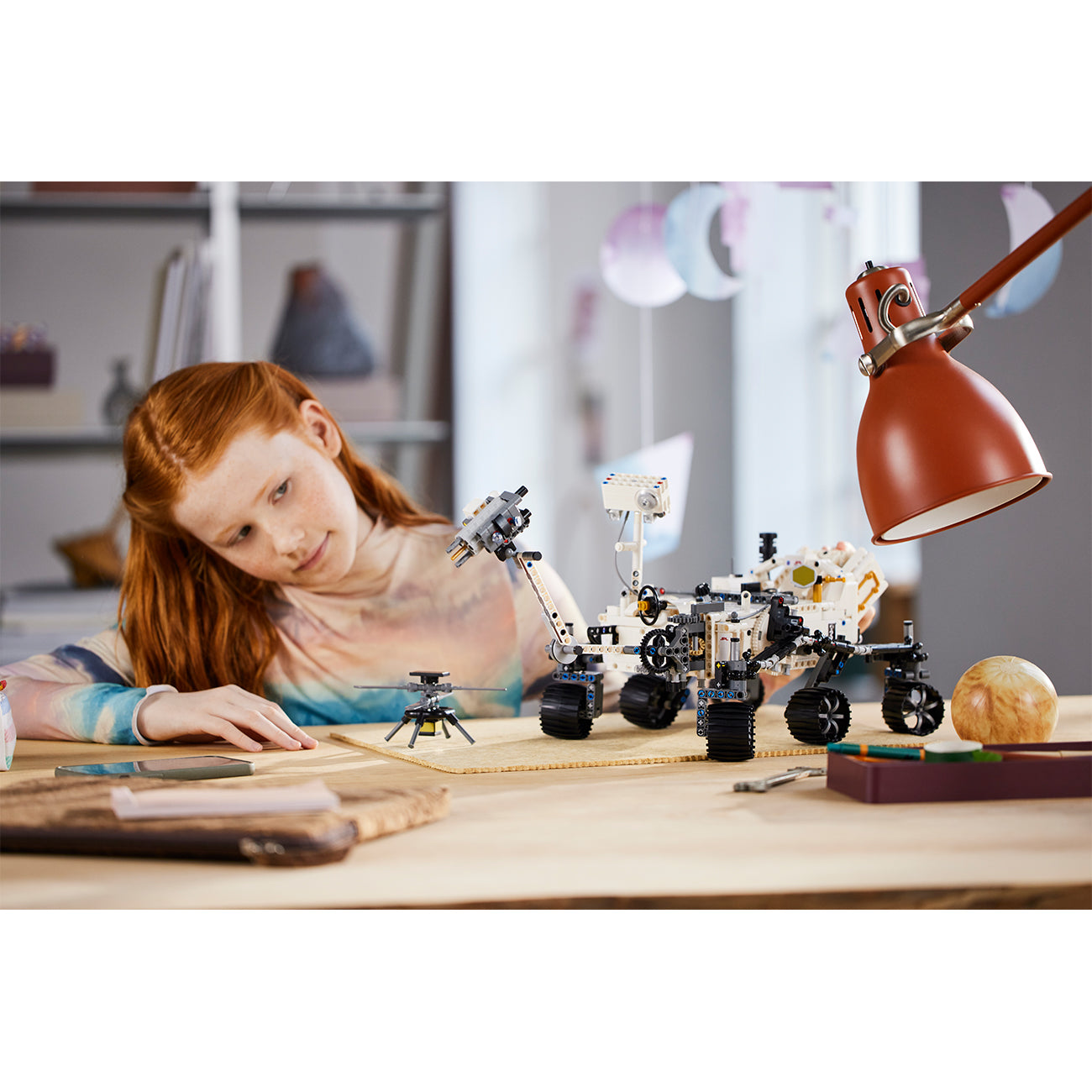 A young girl with red hair sits at a desk, writing in a notebook while studying the LEGO® NASA Mars Rover Perseverance by LEGO, surrounded by books and a lamp in her cozy, well-lit room.