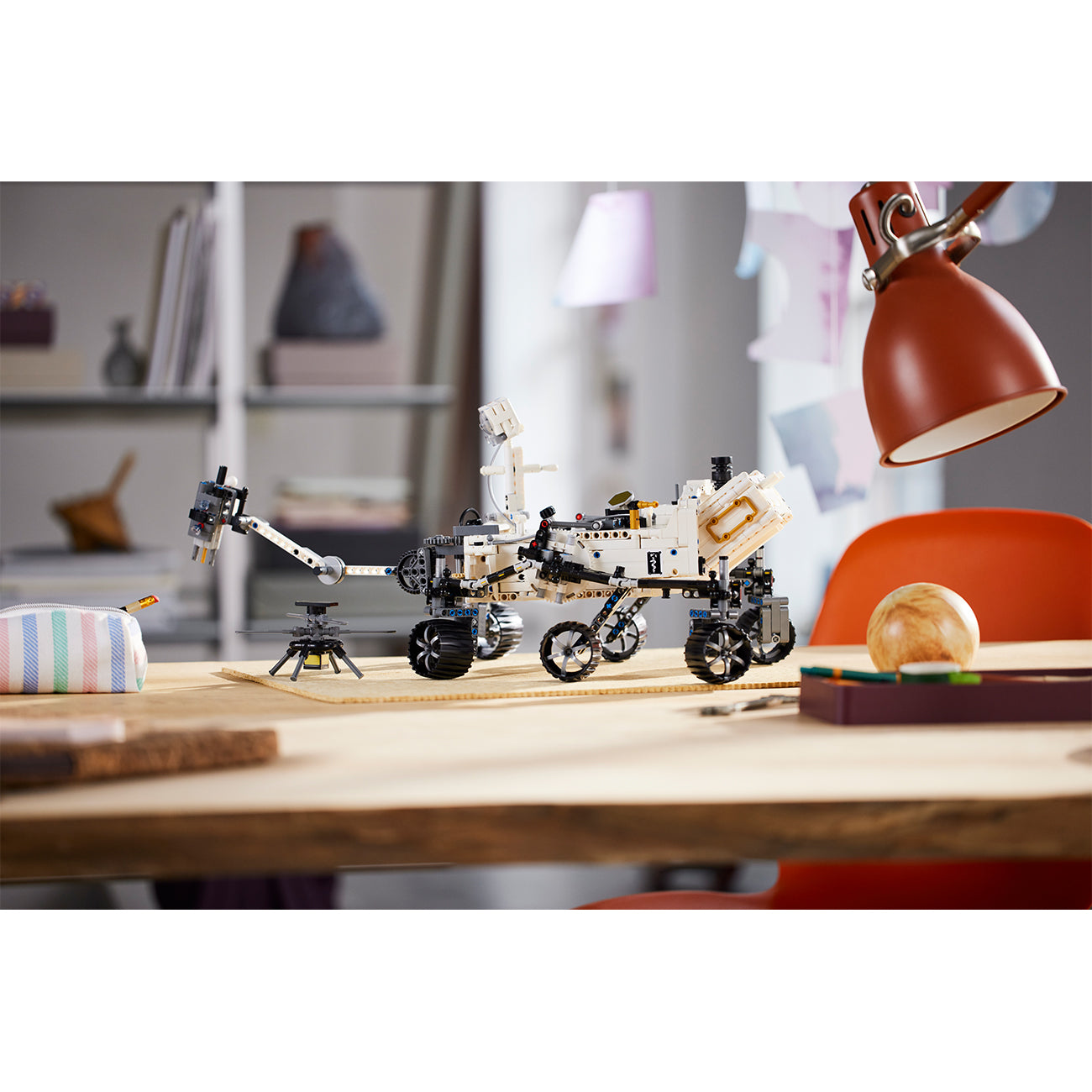 A LEGO® NASA Mars Rover Perseverance by LEGO sits on a wooden table in a study with shelves, a red lamp, books, and a globe—ideal for space exploration play or pairing with an augmented reality app.