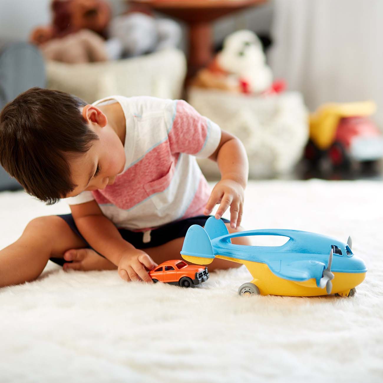 A brown-haired boy, wearing a pink and white striped shirt and shorts, plays on a fluffy white rug with an orange mini car. Hes fascinated by a large blue airplane made from recycled materials. In the background, plush toys and the Green Toys™ Cargo Airplane with Mini Car from The Boeing Store are visible.