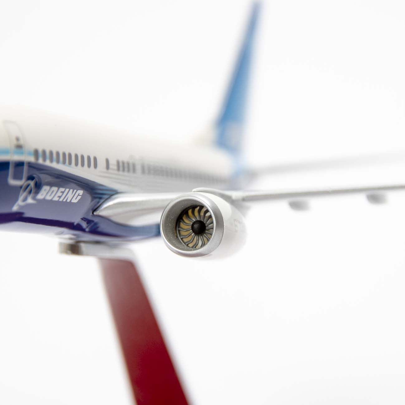 A close-up of The Boeing Stores Boeing Unified 737 MAX 7 1:200 Model showcases a detailed engine. Set against a white backdrop, the 737 MAX 7 logo is visible, with the rudder and blue tail slightly blurred in the distance, emphasizing its renowned fuel efficiency.