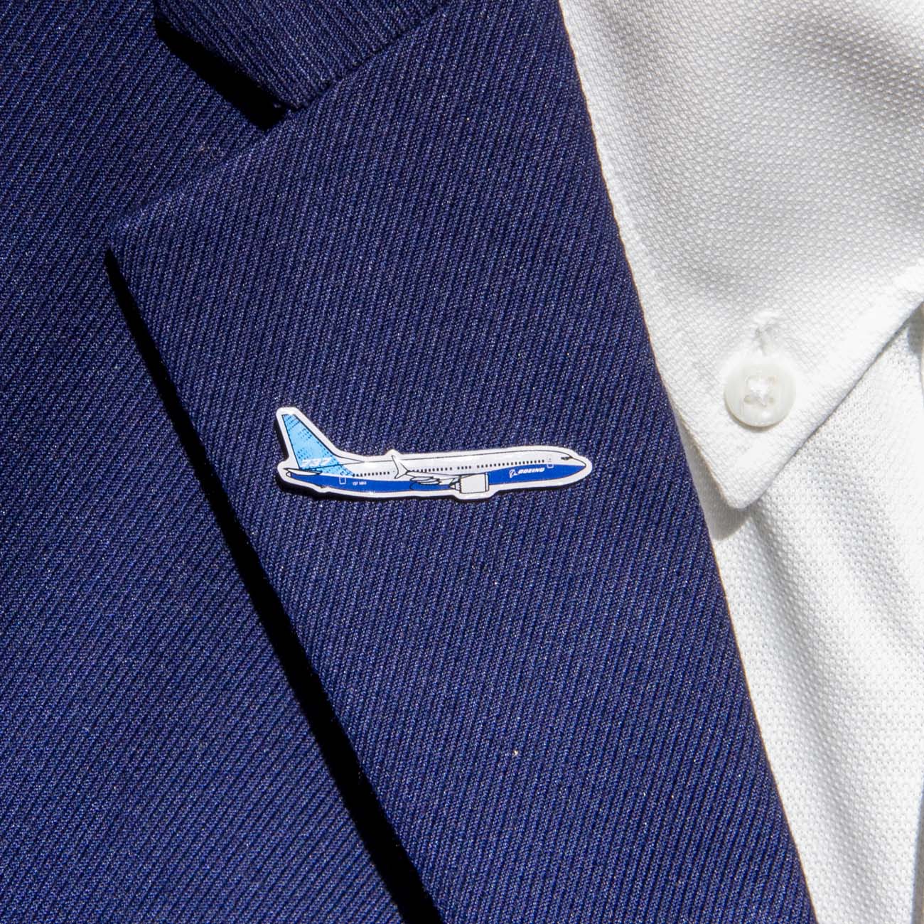 A silver tone lapel pin on the dark blue suit features an intricately designed blue and white aircraft, resembling the Boeing logo. The Boeing Illustrated 737 MAX Lapel Pin by The Boeing Store perfectly complements the imported textured white shirt with crisply visible buttons.