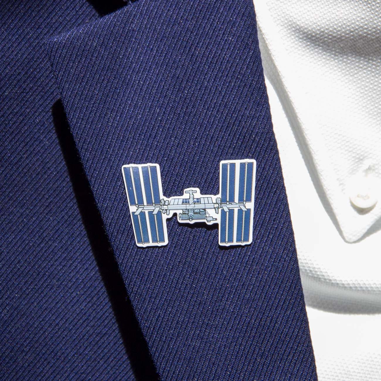 A person wearing a blue pinstripe suit jacket and white shirt showcases The Boeing Stores Boeing Illustrated ISS Lapel Pin. The silver-tone pin, featuring enamel details and an epoxy finish, adds sophistication to any outfit.