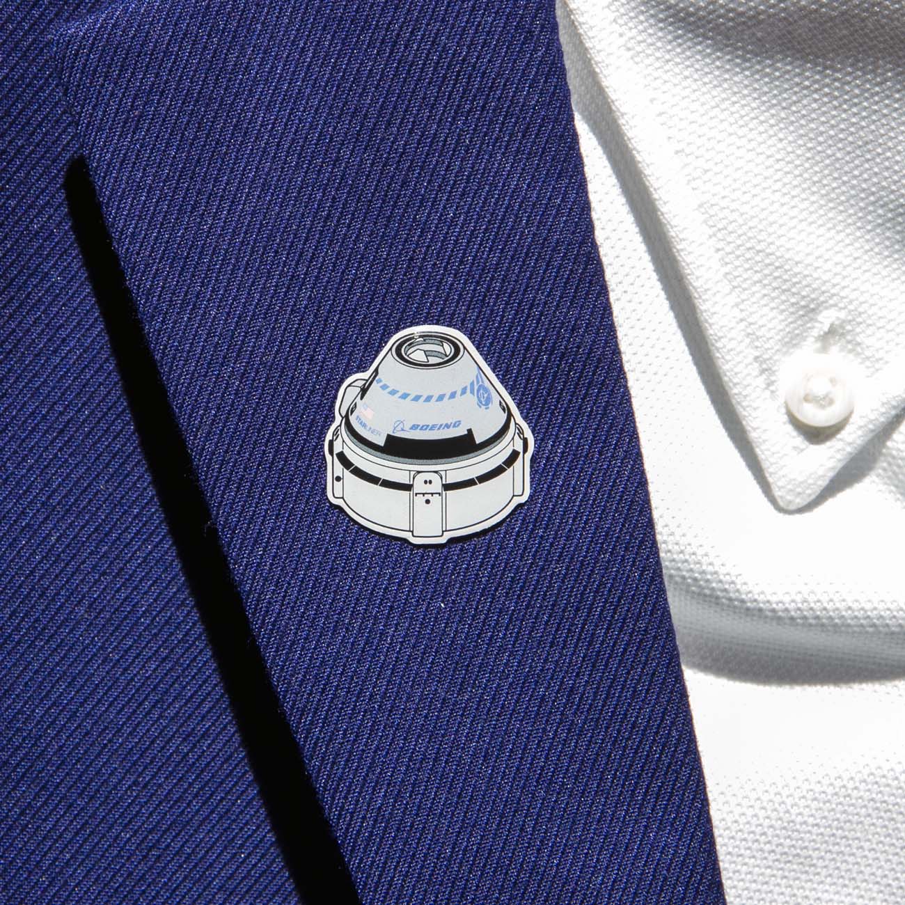 A Boeing Illustrated Starliner Lapel Pin with an epoxy finish and a small logo from The Boeing Store is attached to a blue suit jacket worn over a white textured shirt with a button showing.