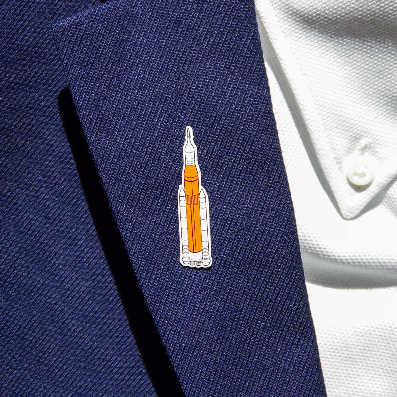 A person in a dark blue suit and white shirt wears a silver-tone lapel pin from The Boeing Store, featuring an orange and white rocket shape, subtly showcasing their connection to aviation.