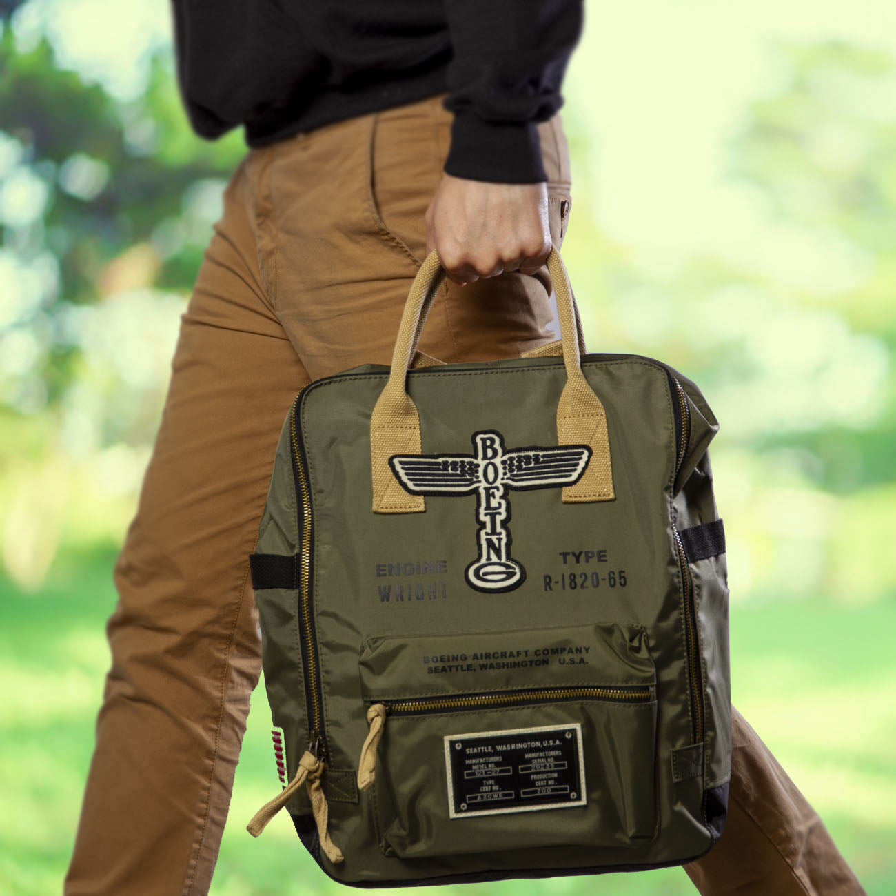 A person strolls through a lush, blurred landscape wearing tan pants and a dark top, carrying The Boeing Stores Red Canoe Backpack, which features aviation-themed prints like propellers and the vintage Boeing airplane logo.