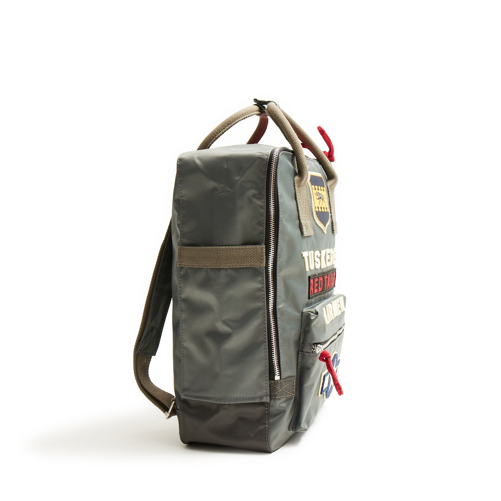 Side view of The Boeing Stores Red Canoe Boeing Tuskegee Red Tails Backpack, made from durable nylon with brown adjustable straps and red handles. Its adorned with vibrant red, blue, and yellow patches and text. This rectangular backpack features a front pocket and zippered compartments for organization.