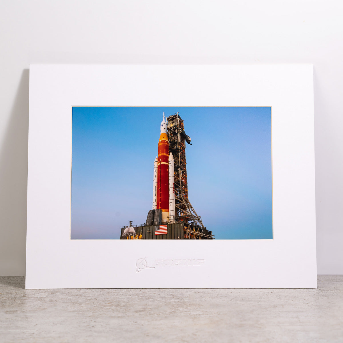 The Boeing Stores Boeing Space Launch System matted print features a red and white rocket against a blue sky with an attached launch tower, framed in white and embossed with the Boeing logo. It showcases high-resolution digital printing and rests elegantly on a gray surface.