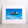 The Boeing Store presents the Boeing F/A-18 Super Hornet Matted Print - Small, showcasing a fighter jet gliding through a clear sky with clouds. The Boeing logo is embossed on the white frame, and the high-resolution digital print rests elegantly on a light-colored surface.