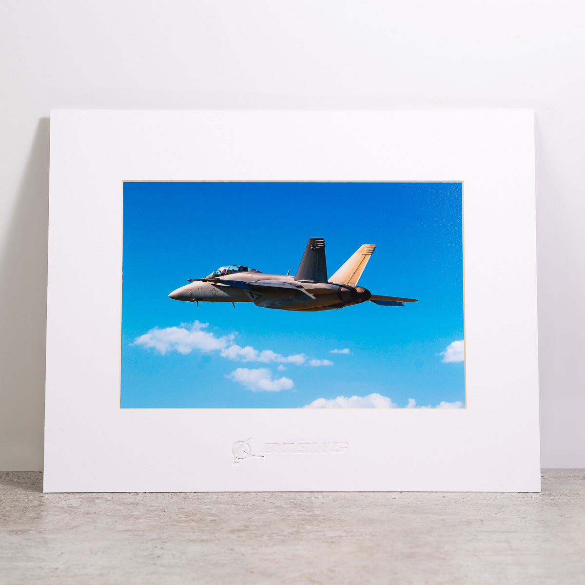 The Boeing Store presents the Boeing F/A-18 Super Hornet Matted Print - Small, showcasing a fighter jet gliding through a clear sky with clouds. The Boeing logo is embossed on the white frame, and the high-resolution digital print rests elegantly on a light-colored surface.