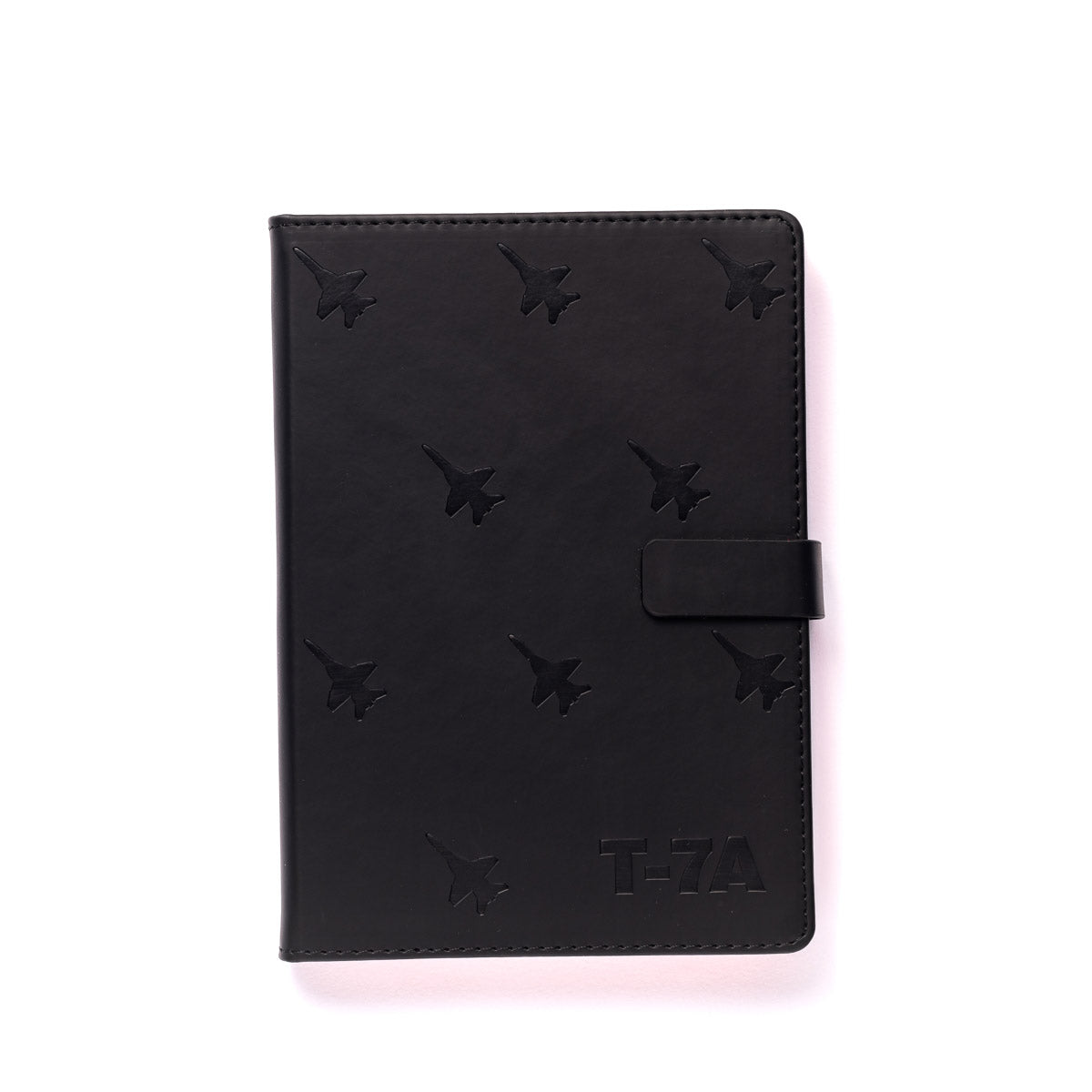 The Boeing Stores Boeing T-7A Red Hawk Notebook is a black leather accessory embossed with fighter jet patterns. It features a strap closure and T-7A text in the corner, elegantly photographed against a plain, light background, exuding an aura of aviation artistry.