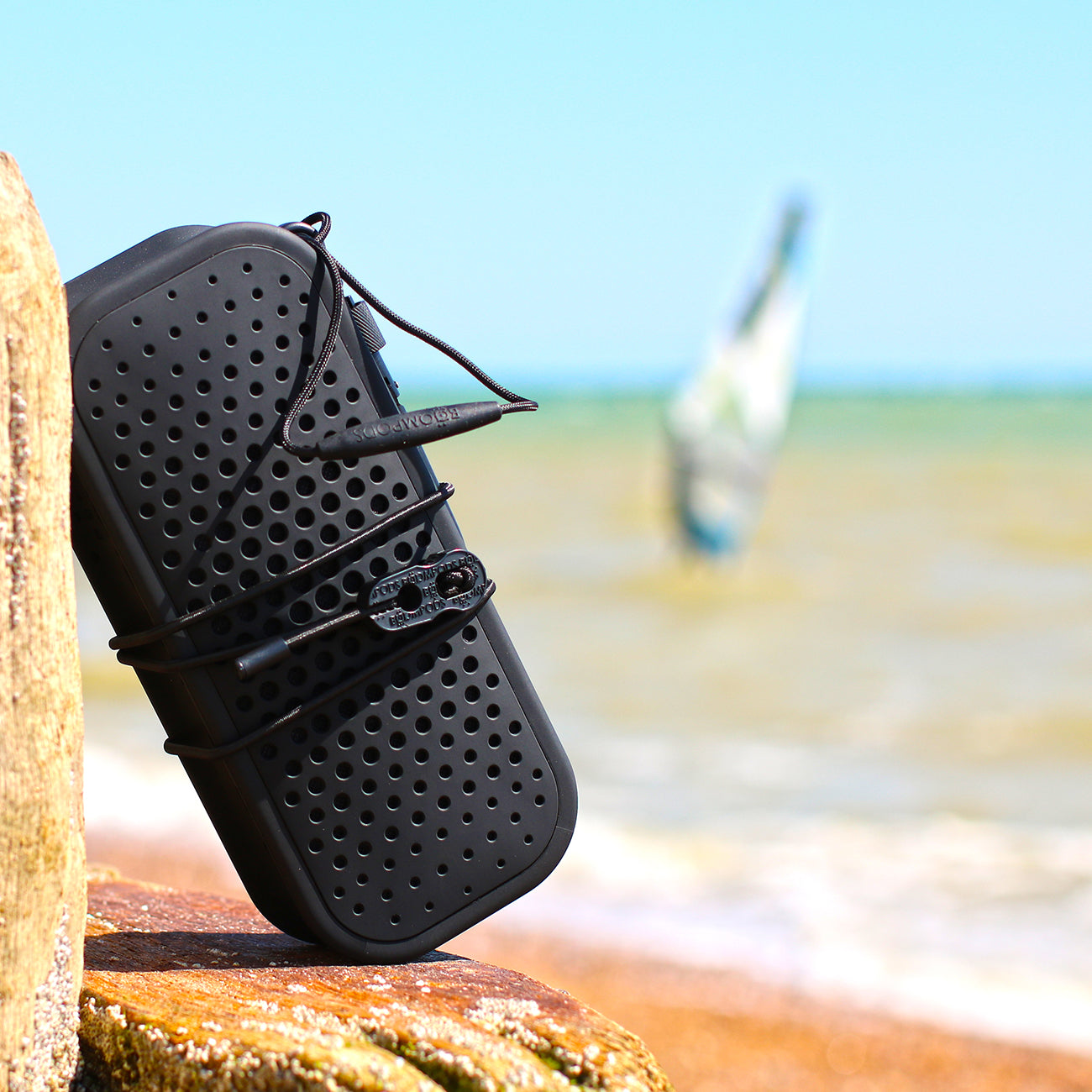 A black Boompods Blockblaster Speaker from The Boeing Store, featuring a perforated design, sits on a beach rock. The ocean and a windsurfer blur in the background under a clear blue sky, promising an immersive audio experience.