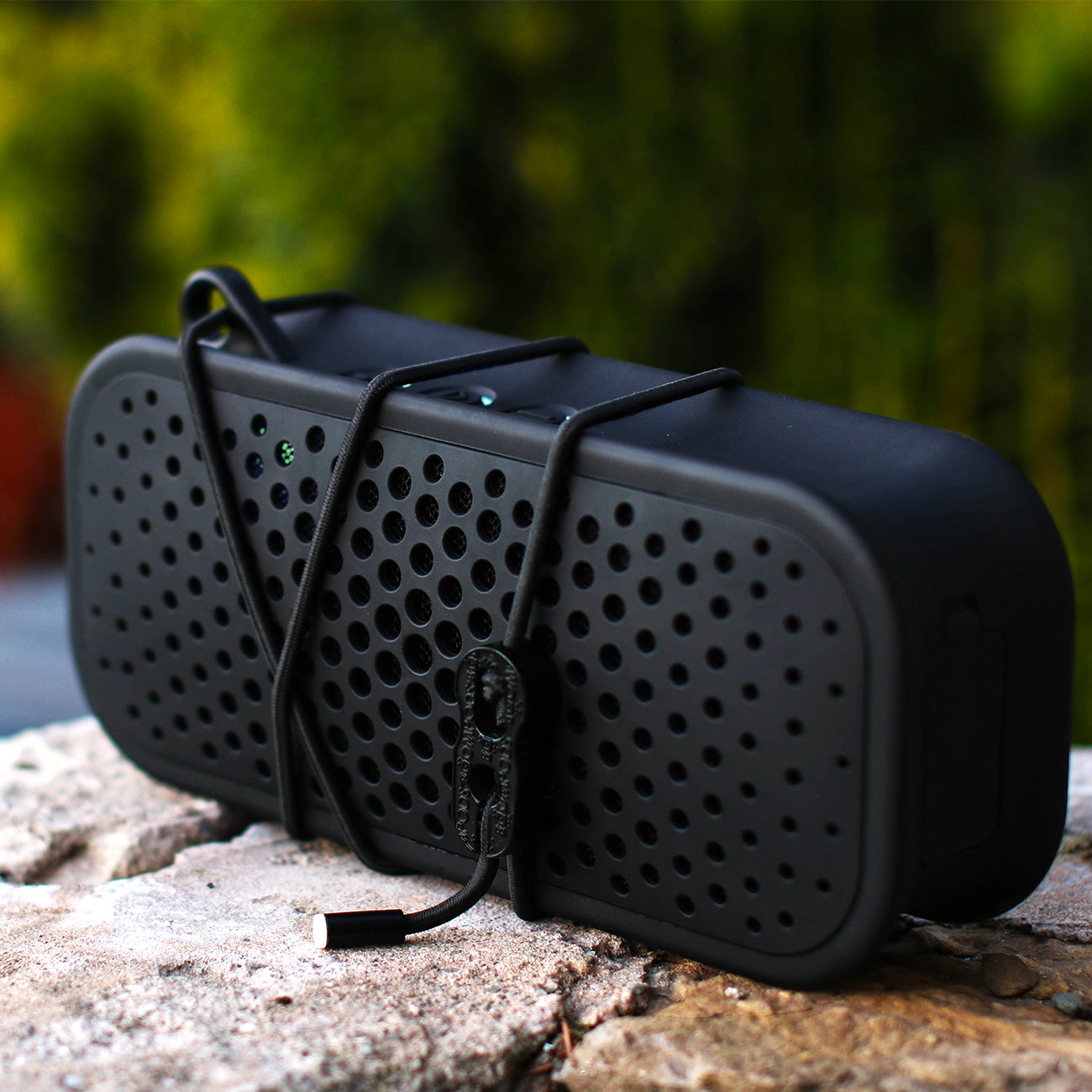 A black Boompods Blockblaster Speaker by The Boeing Store, featuring a perforated design and secured with an elastic cord, rests on stone. It delivers Hi-Fi wireless sound with impressive clarity and rich bass against a blurred backdrop of lush greenery.
