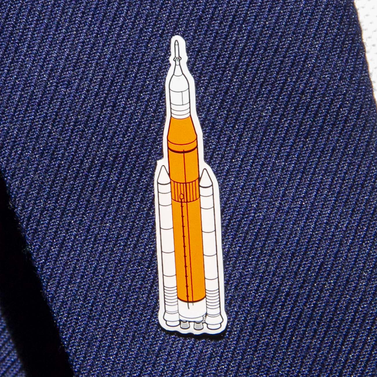 The Boeing Illustrated SLS Lapel Pin features a rocket with an orange body and white boosters on a textured dark blue fabric, reminiscent of space-inspired artwork, enhanced with silver tone sophistication.
