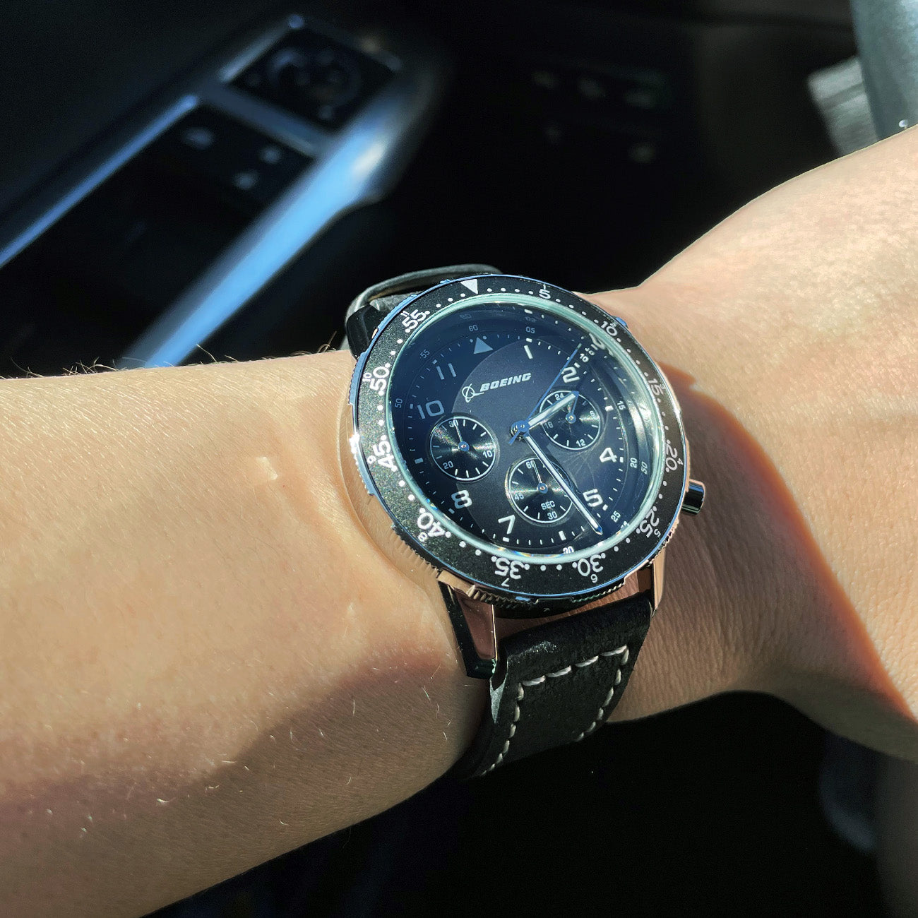 A wrist sports a Boeing Black Leather Watch: round face, silver markings, iconic Boeing logo. It features a stainless steel case and black leather strap with white stitching against a car interior backdrop. Available at The Boeing Store.