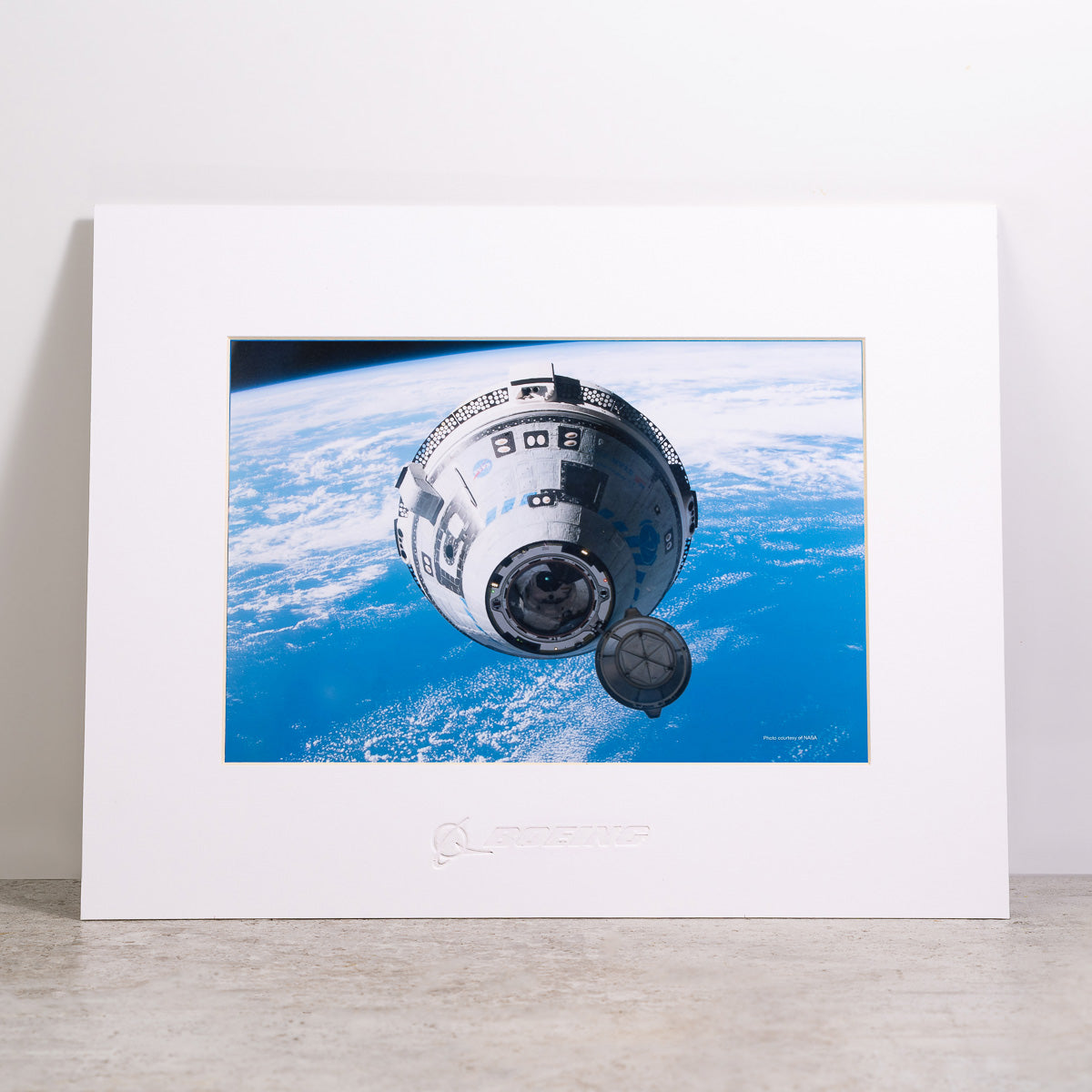 The Boeing CST-100 Starliner Matted Print - Small from The Boeing Store features a photo of the spacecraft orbiting Earth, displayed in a white matted frame with a Boeing logo on the bottom, set against a plain background.