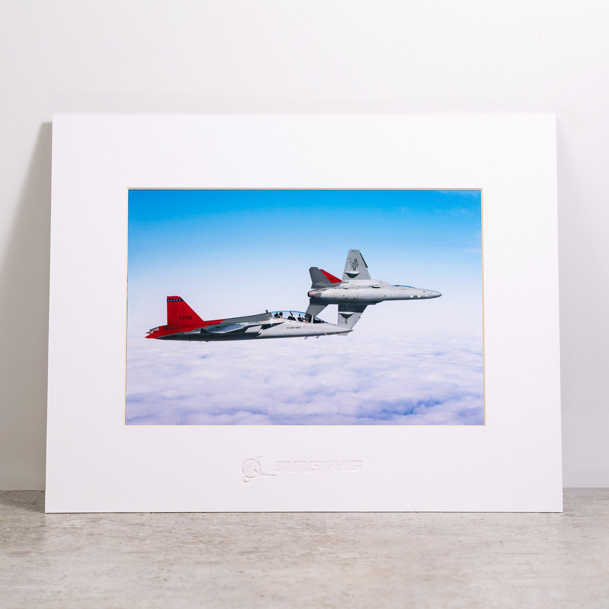 The Boeing Store’s T-7A Red Hawk Matted Print (Small) features two military jets—one with a red tail, one gray—soaring above the clouds, set against a white wall. The Boeing logo highlights its quality digital printing.